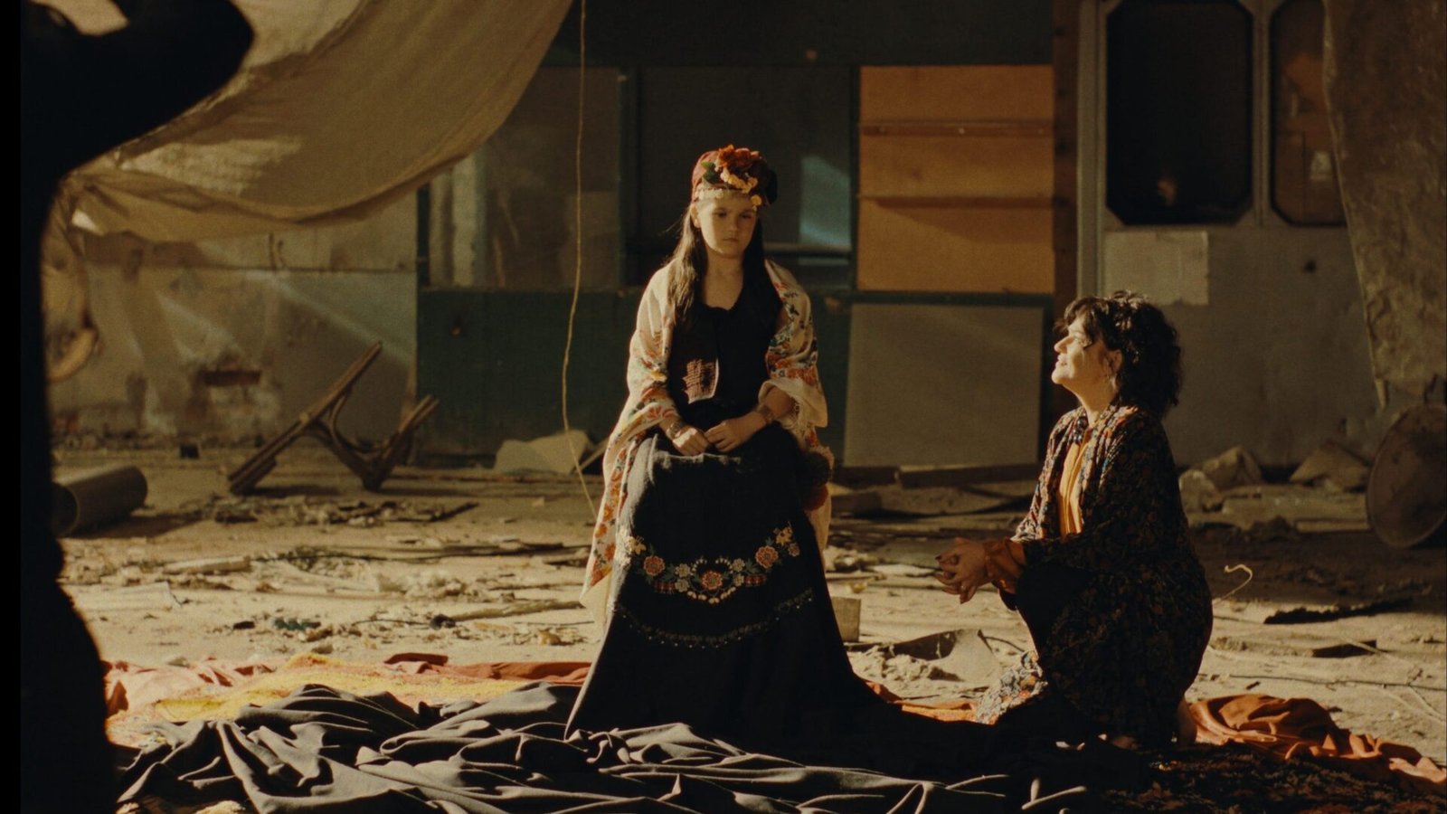 Two women in a ruined hall, one in a traditional embroidered dress standing over the other, who kneels on draped fabric