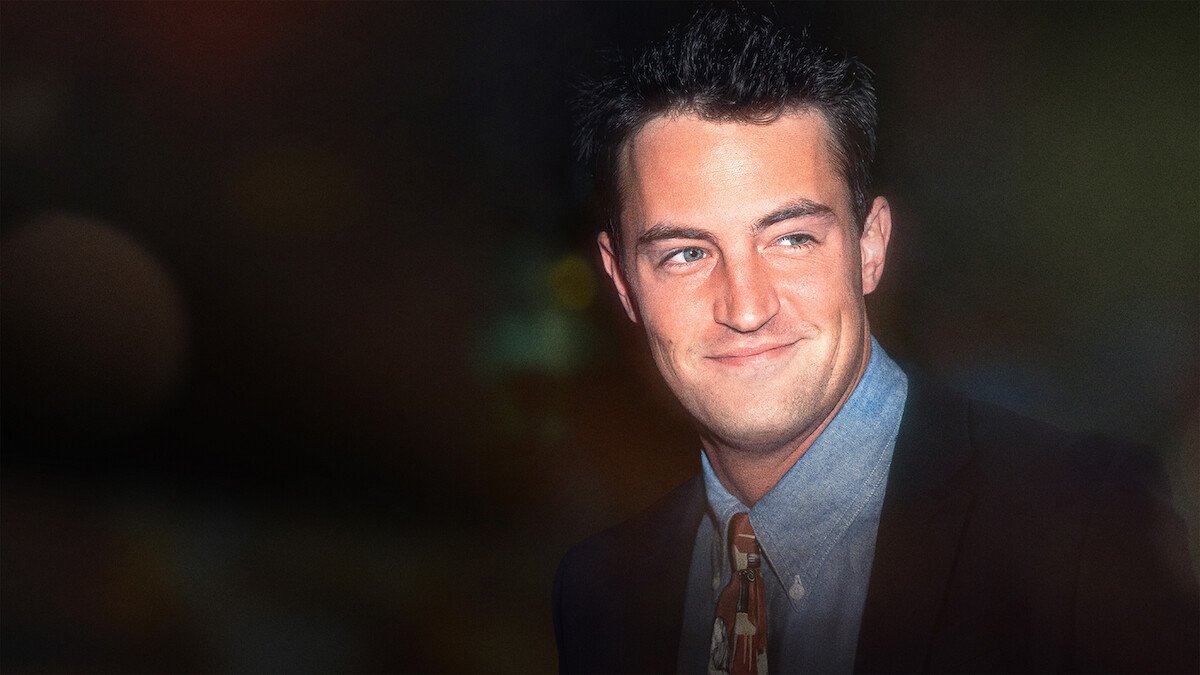 Matthew Perry in a suit and patterned tie, smiling against a dark background