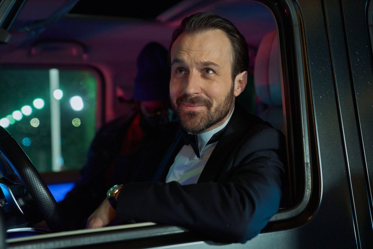 A man in a suit sits in the driver's seat of a car at night, smiling out of the window, city lights behind him, in Lupin Part 4.